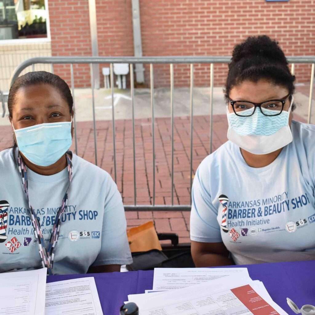 Two women in gray shirts with blue face masks sit outside behind a purple table with papers on it.
